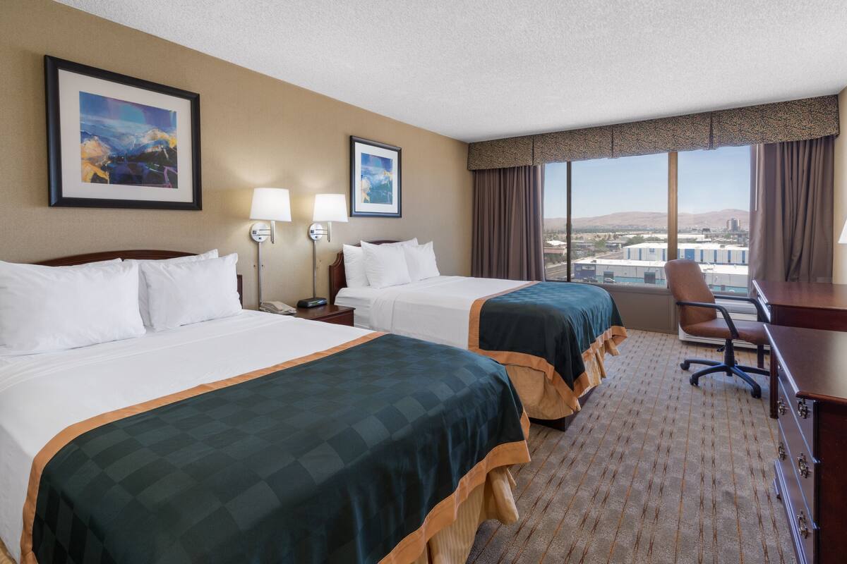 A hotel room with two beds, a desk, and a chair. There are two framed pictures on the wall, a window with a view of buildings, and a carpeted floor.