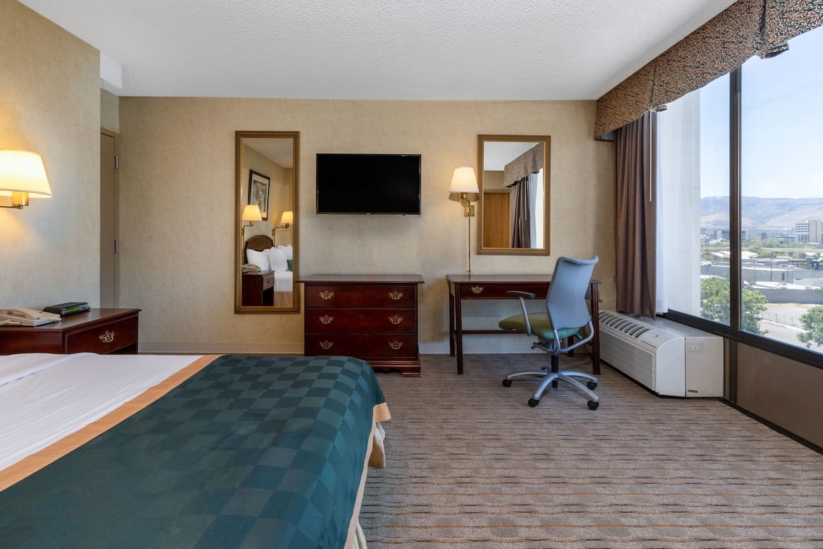A hotel room with a bed, a desk, a chair, a TV, a dresser, and a large window with a city view.