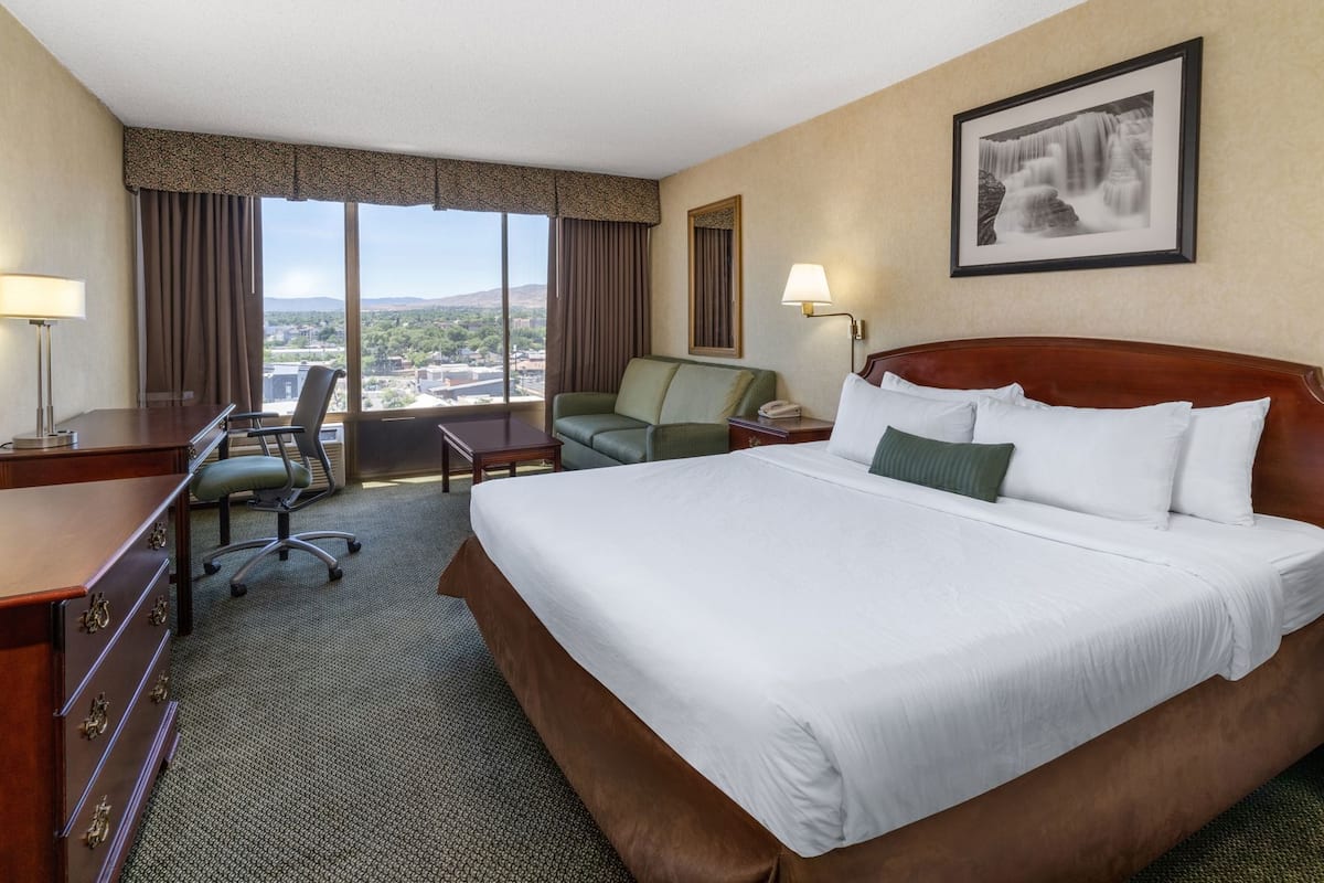 A hotel room with a large bed, a desk with a chair, a small table, a green armchair, and a view of the cityscape through large windows.