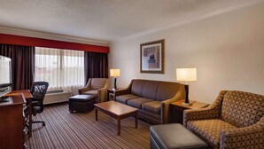 A hotel room with a sofa, armchair, ottoman, coffee table, TV, and air conditioning unit. - SureStay by Best Western Miles City (Miles City)