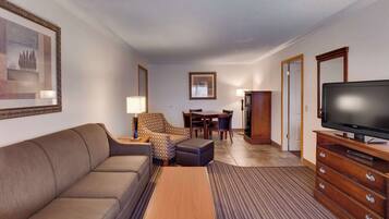 A hotel room with a sofa, patterned armchair, dining table with chairs, a television, and a cabinet.