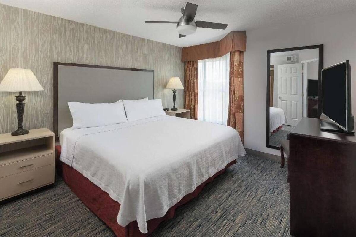 Une chambre d’hôtel comprenant un lit, deux tables de chevet avec des lampes, une télévision, un ventilateur de plafond et un miroir.
