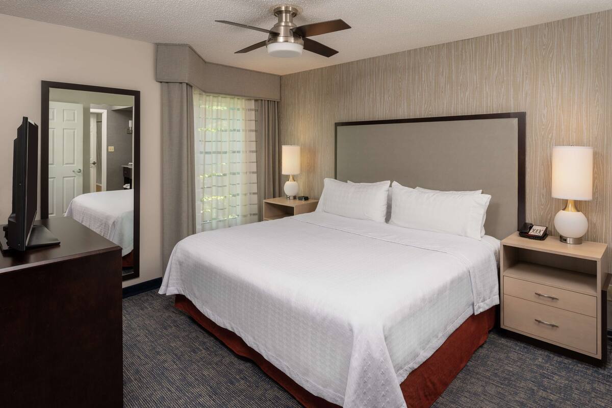 Une chambre d’hôtel avec un grand lit, deux tables de chevet avec des lampes, un ventilateur de plafond, un miroir et un téléviseur posé sur une commode.