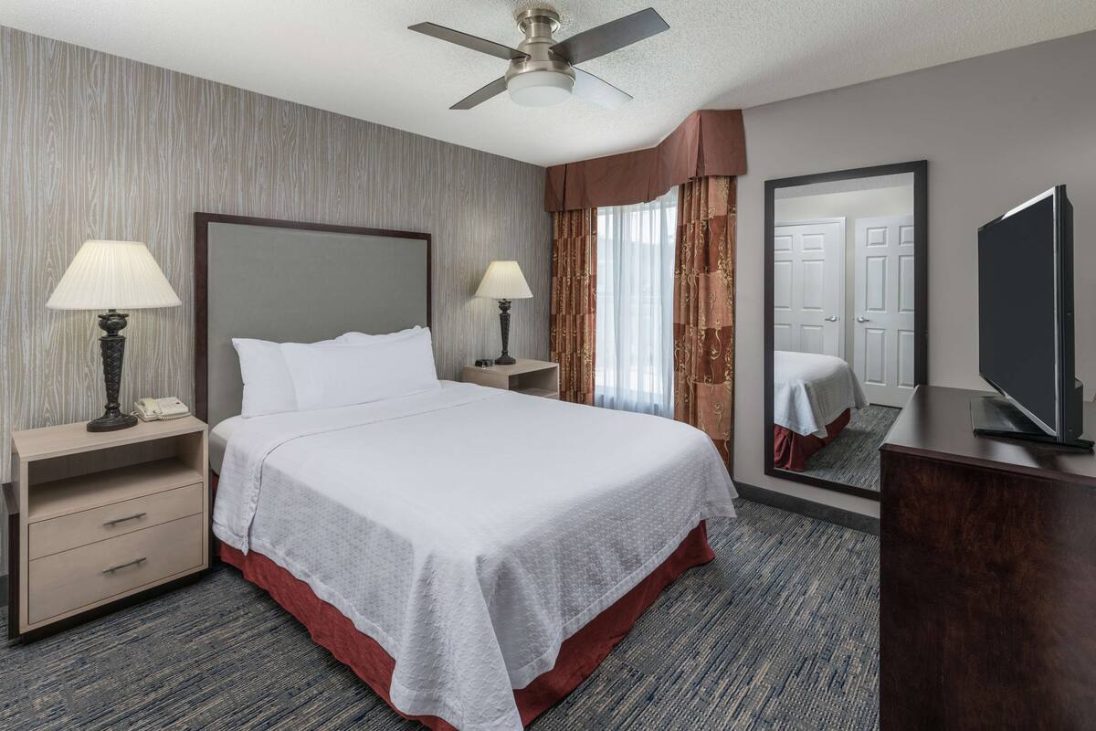 Une chambre d’hôtel comprenant un lit, deux tables de chevet, des lampes, un ventilateur de plafond, une télévision et un miroir.