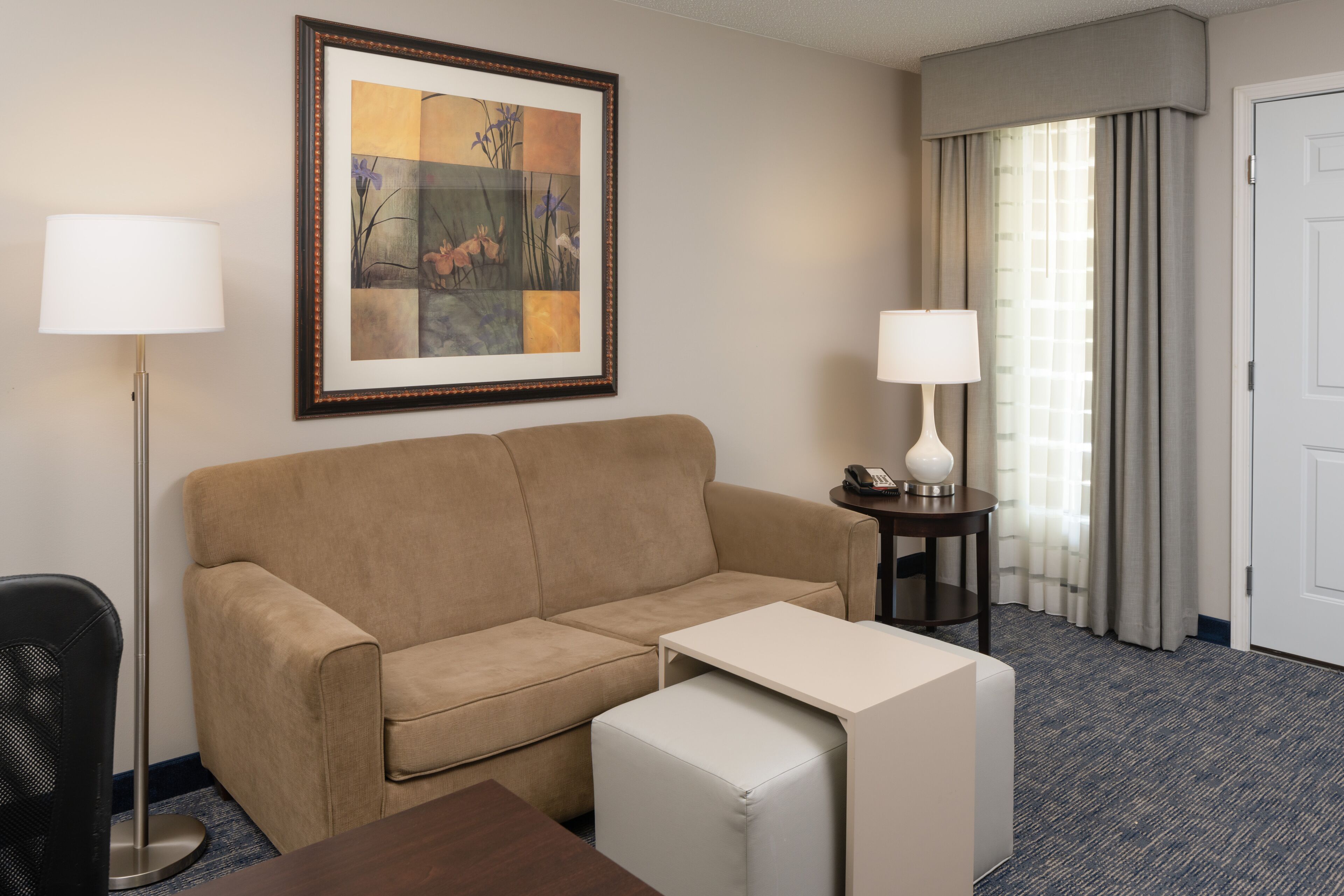 Habitación de hotel con un sofá color crema, una pequeña mesa blanca, una lámpara de pie y un lámpara de pie adicional, además de una obra de arte enmarcada en la pared.