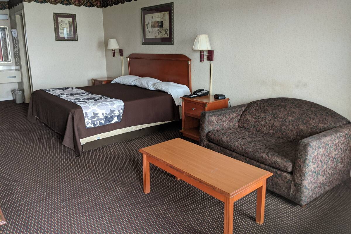 A hotel room with a bed, a wooden nightstand with a lamp, a floral-patterned armchair, and a small wooden table.