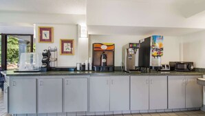 A kitchen area with a coffee machine, a microwave, and a juice dispenser. There are framed pictures on the wall and a window with a view of trees. - Econo Lodge Garner - Raleigh (Garner)
