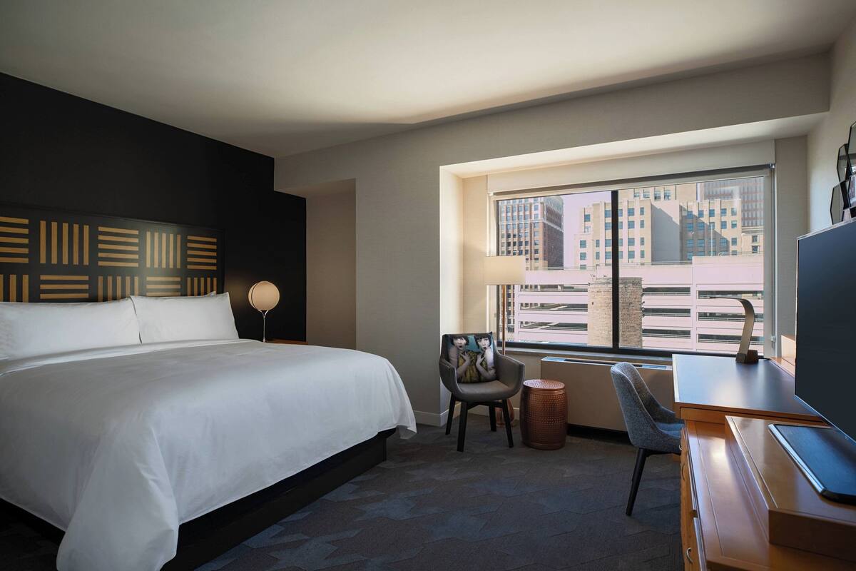 A hotel room with a large bed, a desk, a chair, a lamp, and a view of buildings through the window.