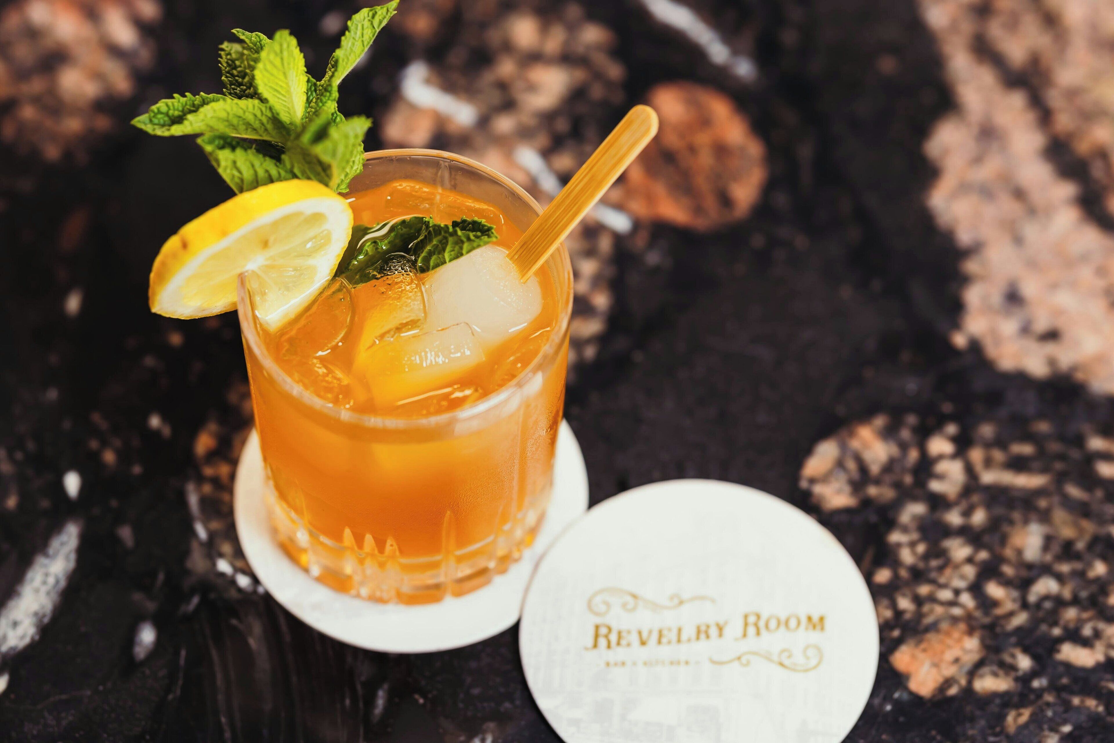 A cocktail with mint and lemon garnish, served in a glass with a straw, on a coaster with "Revelry Room" text.