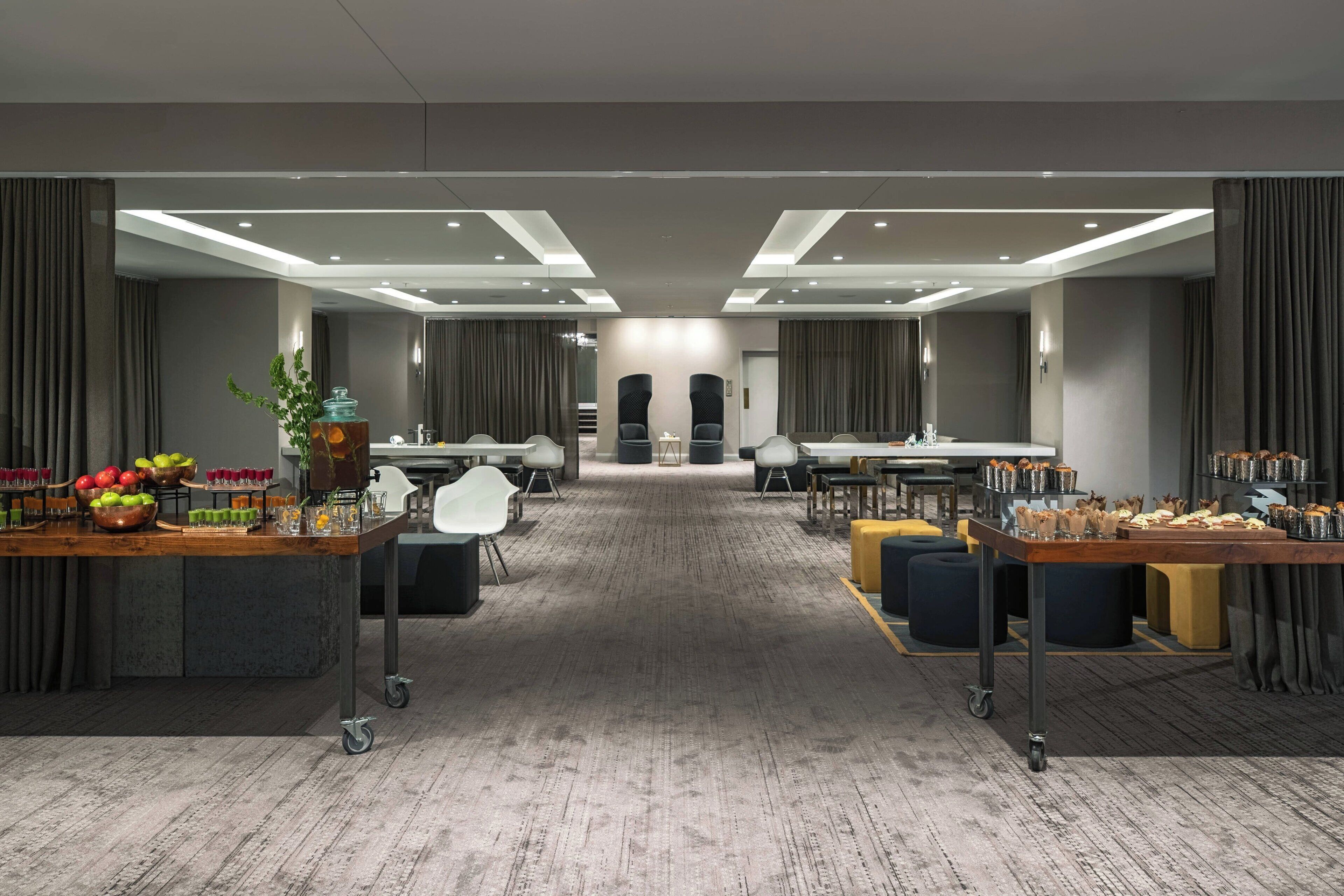 A spacious hotel room with a large dining area, a buffet table with food and drinks, and a seating area with chairs and ottomans.