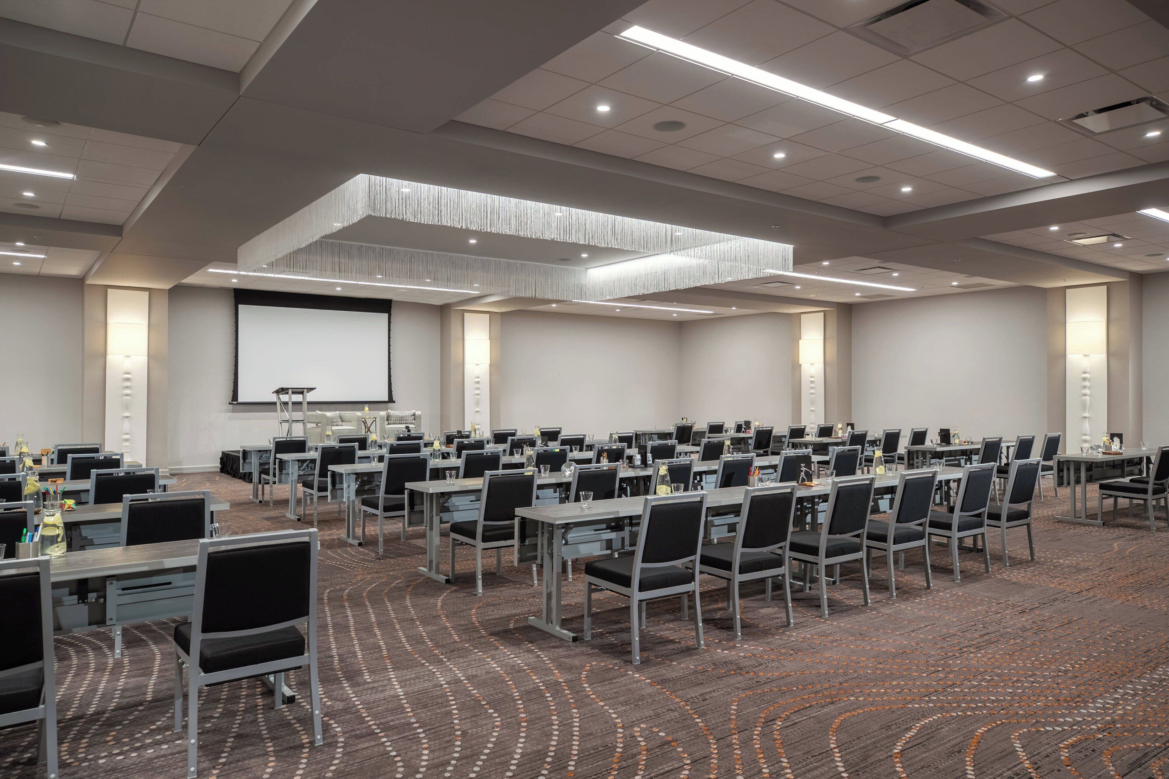 A conference room with multiple tables and chairs, a projector screen, and a carpeted floor.