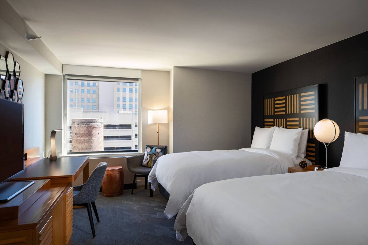 A hotel room with two beds, a desk, a chair, and a window with a city view.