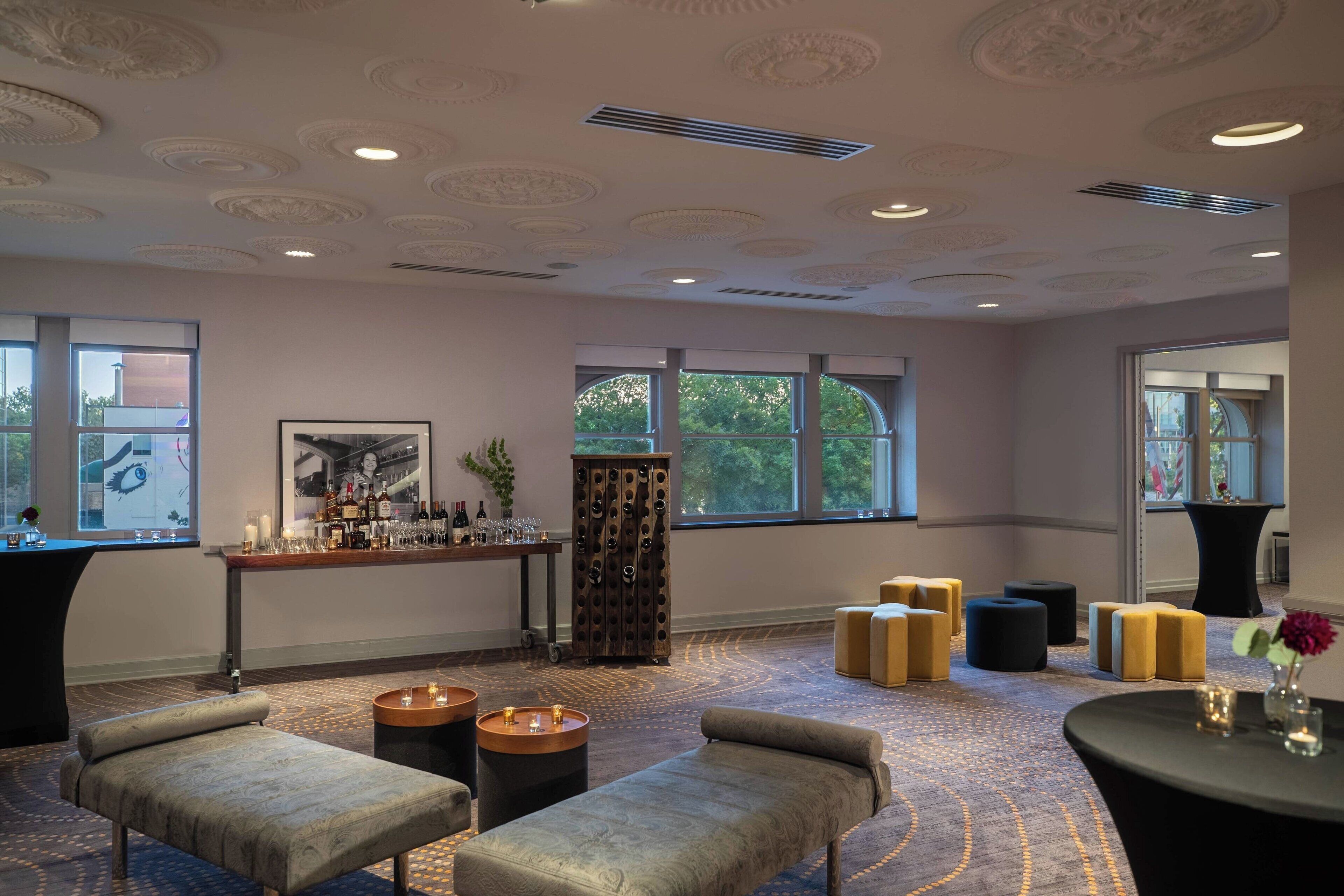 A spacious room with a bar area, seating with ottomans, and large windows providing natural light.