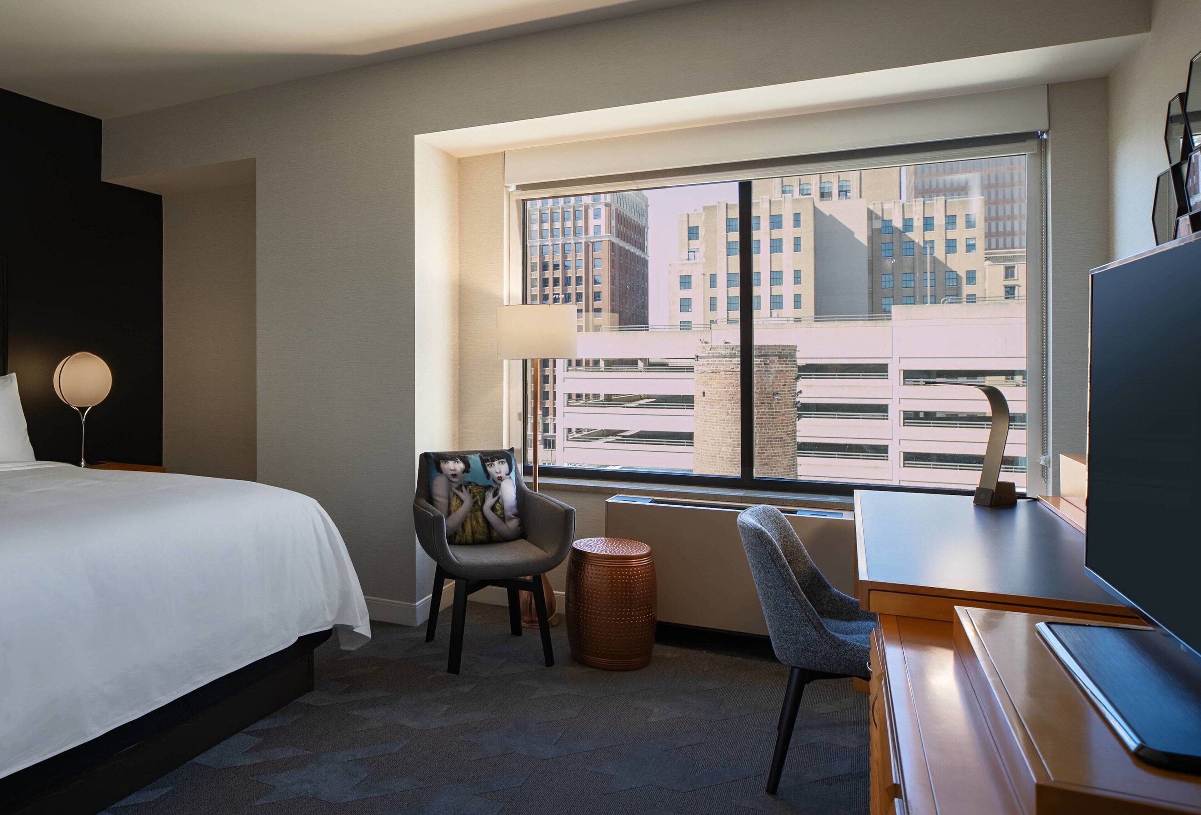 A hotel room with a large bed, a desk, a chair, a lamp, and a view of buildings through a window.