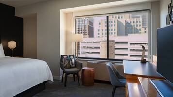 A hotel room with a large bed, a desk, a chair, a lamp, and a view of buildings through a window.
