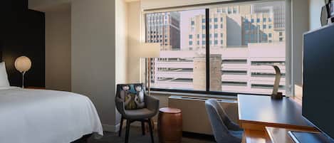 A hotel room with a large bed, a desk, a chair, a lamp, and a view of buildings through a window.