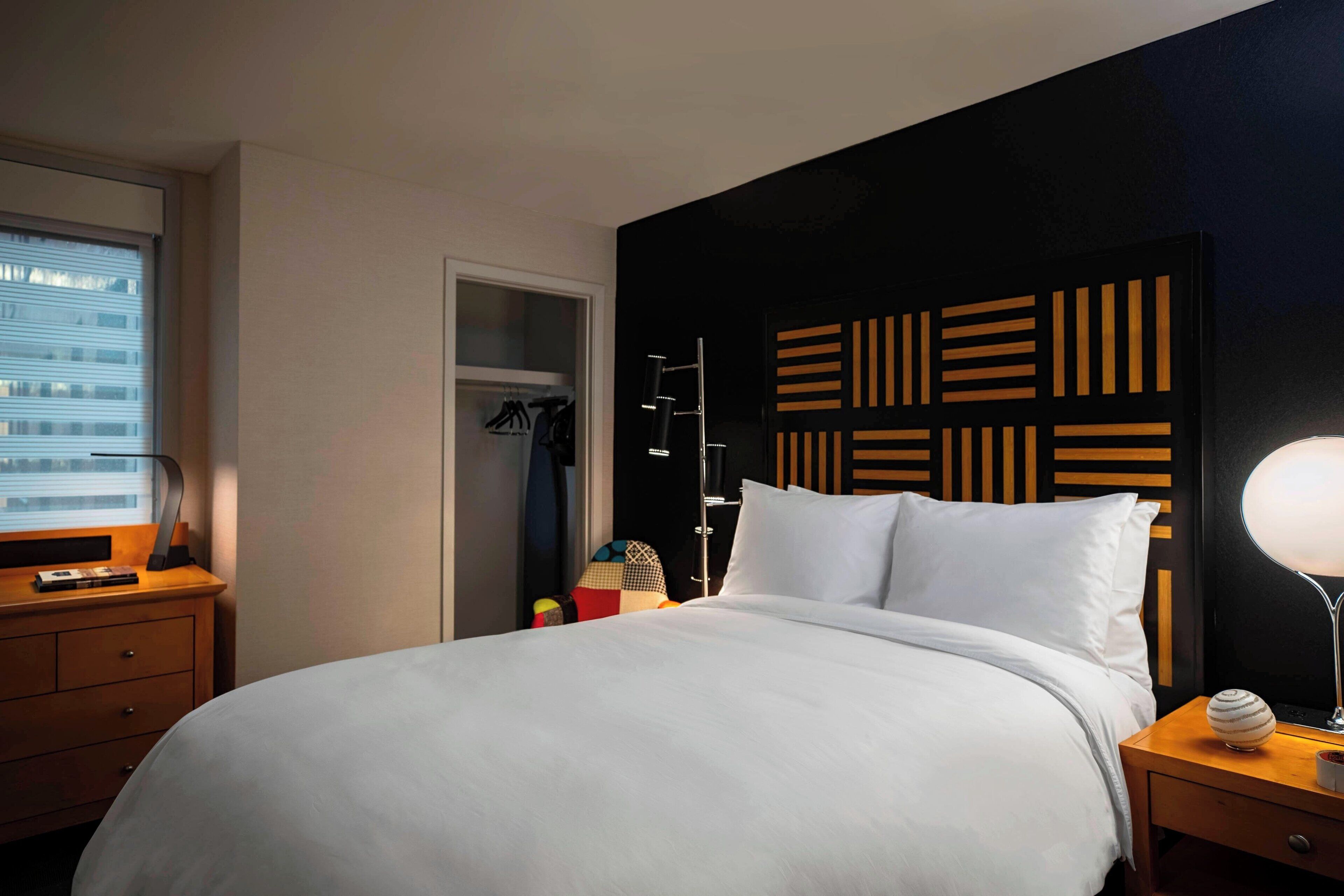 A hotel room with a large bed, a bedside table with a lamp, a wardrobe, and a window with blinds.