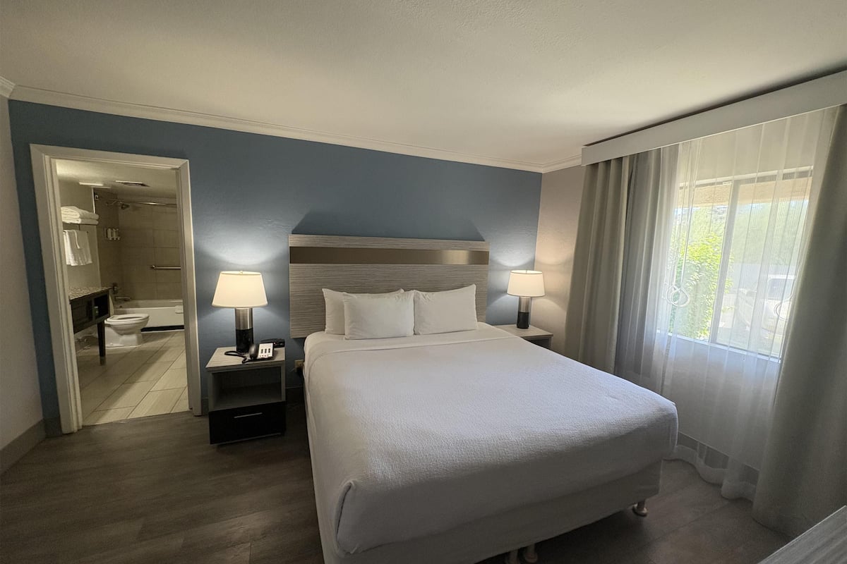 A hotel room with a large bed, two bedside lamps, a nightstand, a bathroom visible through an open door, and a window with curtains.