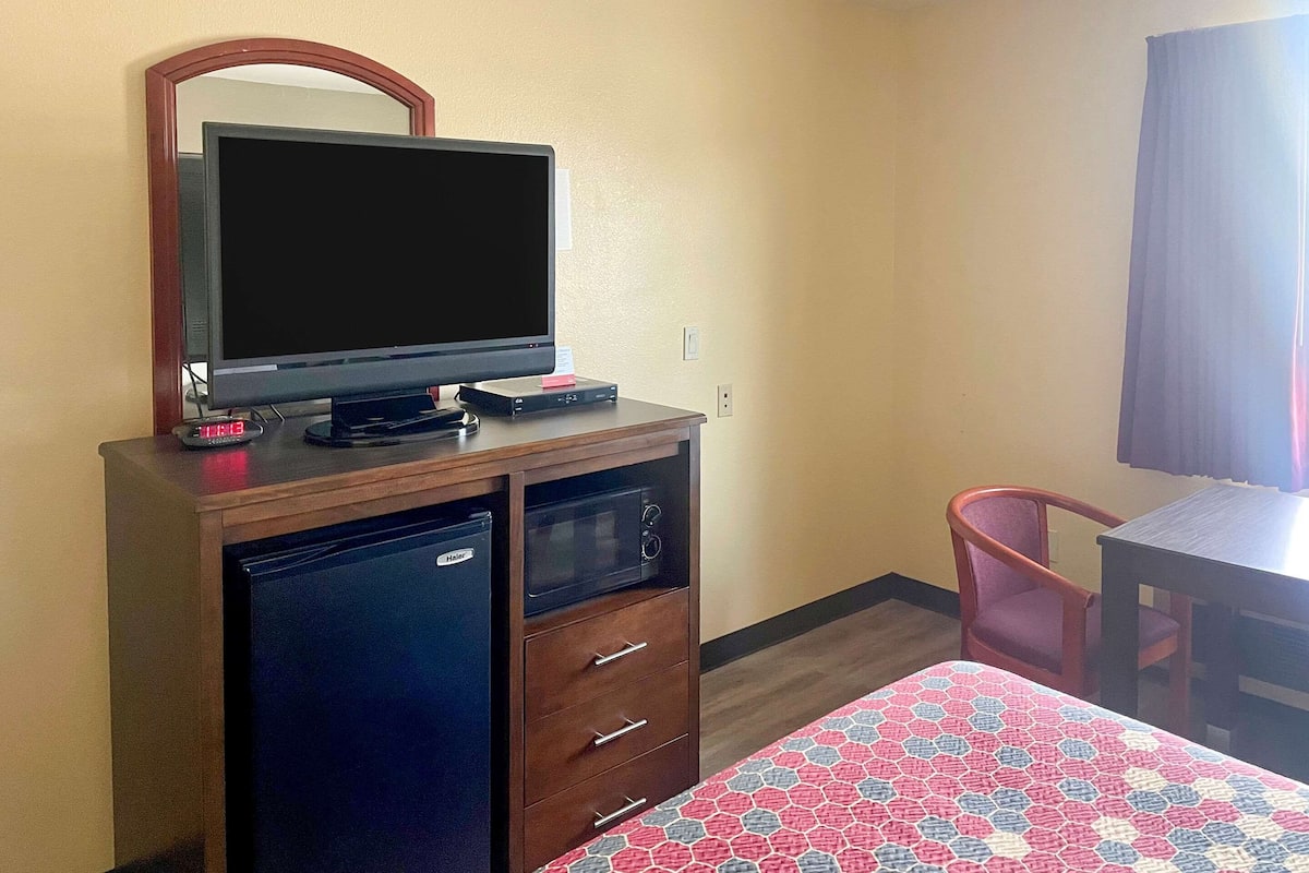 A hotel room with a television on a wooden cabinet, a small refrigerator, a desk, and a bed with a patterned bedspread.