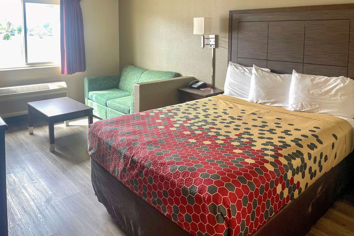 A hotel room with a bed, a desk, a chair, a nightstand, a lamp, and a window with curtains.