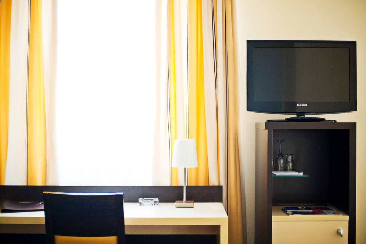 Habitación de hotel con escritorio, televisión, lámpara y un pequeño mueble con objetos decorativos.