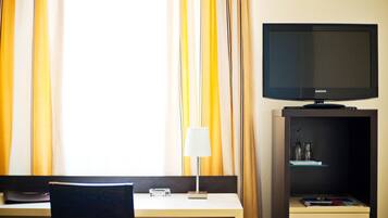Quarto de hotel com escrivaninha, televisão, abajur e um pequeno armário com objetos decorativos.
