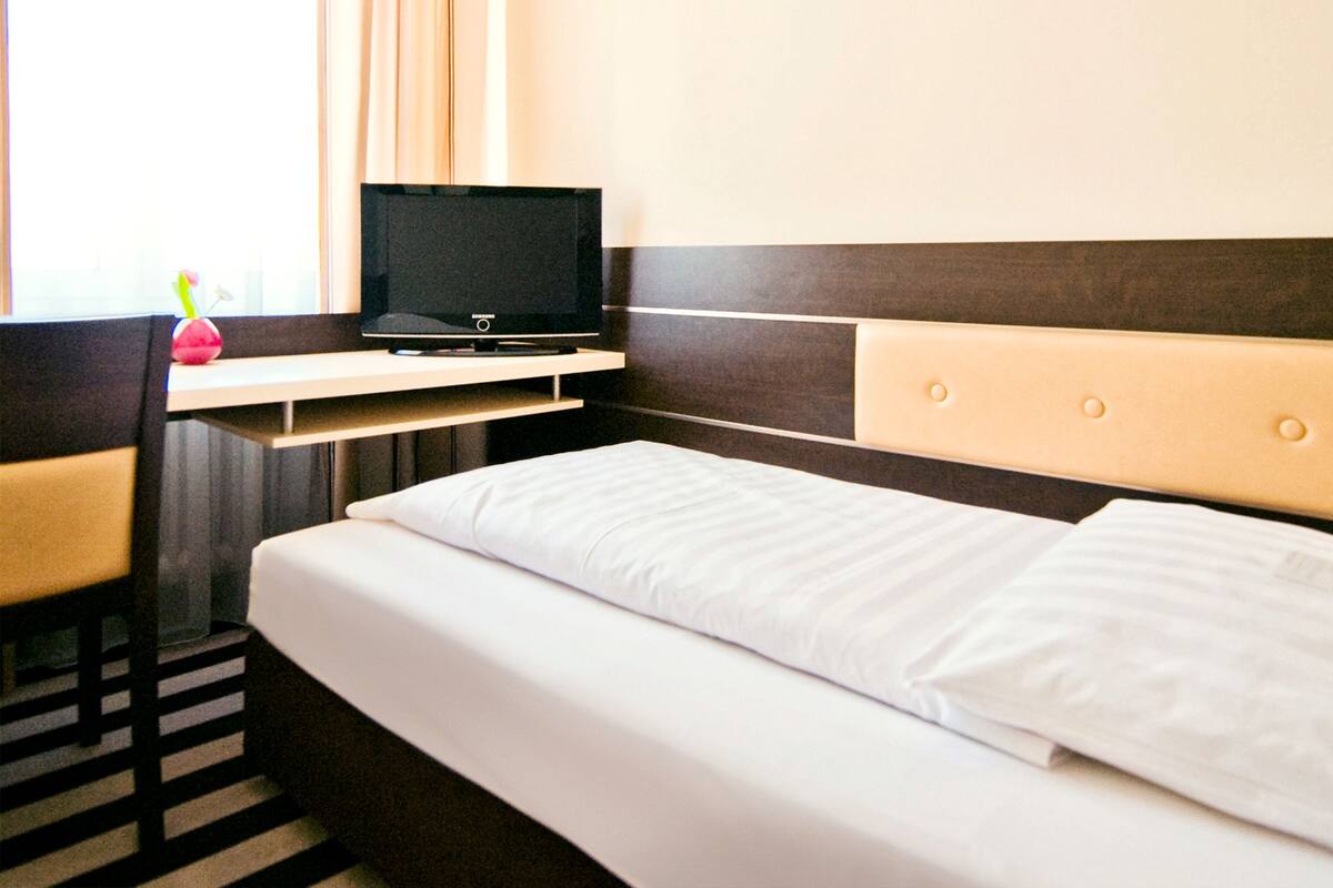 Una habitación de hotel con una cama, un televisor en un soporte, una mesita pequeña y una silla.