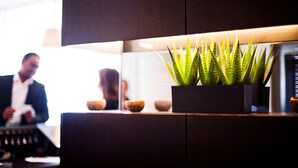 Un mostrador de recepción moderno con una planta en maceta, una caja registradora y una persona desenfocada al fondo.