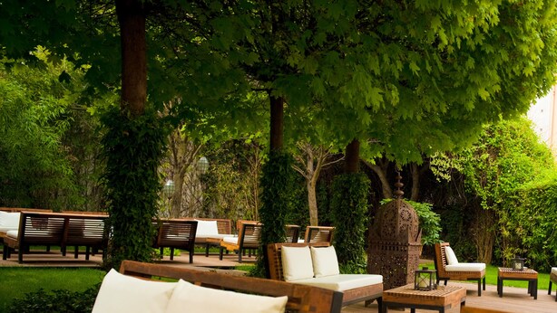 Un espacio al aire libre con mobiliario de madera y cojines blancos, rodeado de exuberante vegetación y árboles.