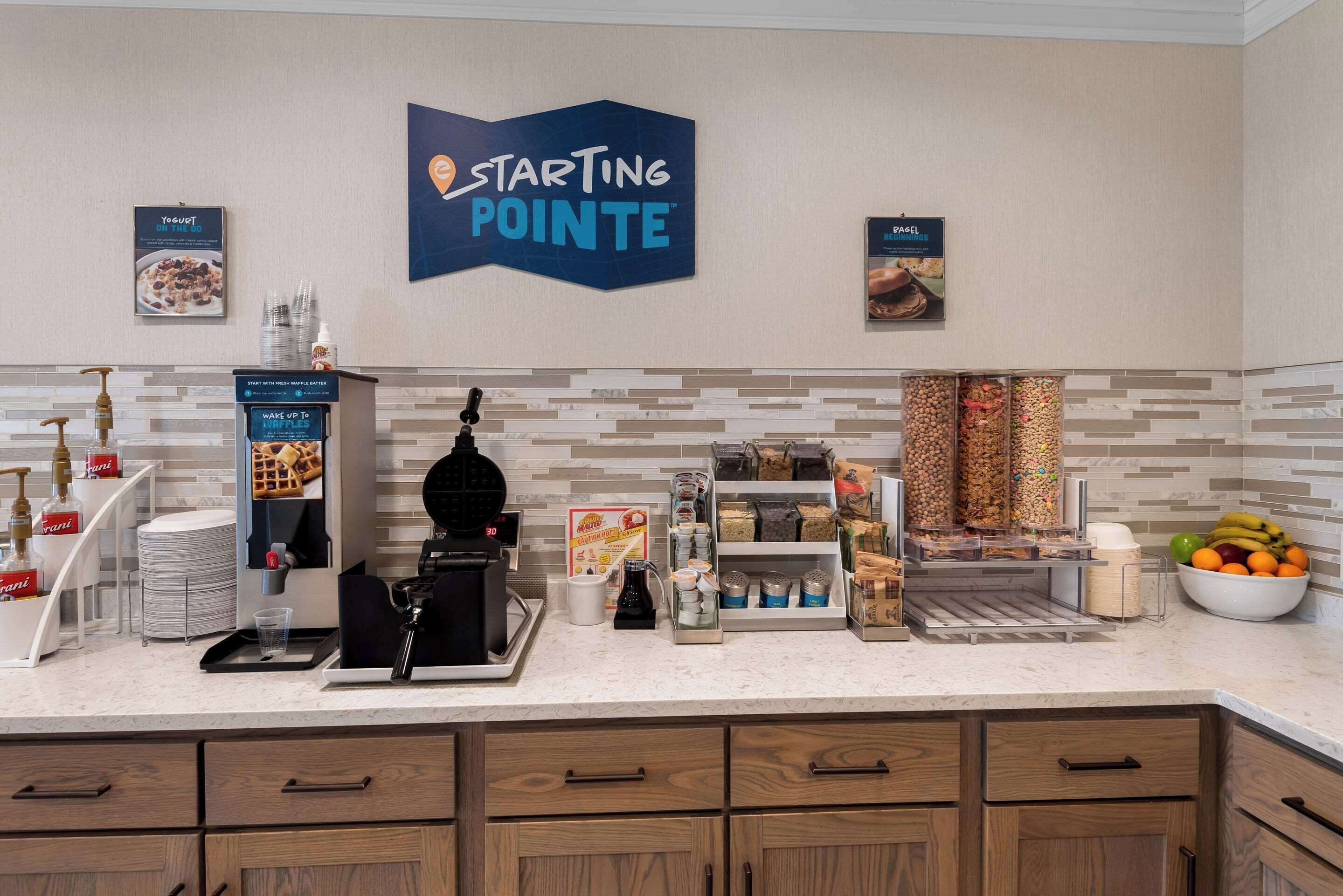 Una kitchenette de hotel con un letrero de Starting Pointe, máquina para hacer gofres, cafetera y diversos alimentos.