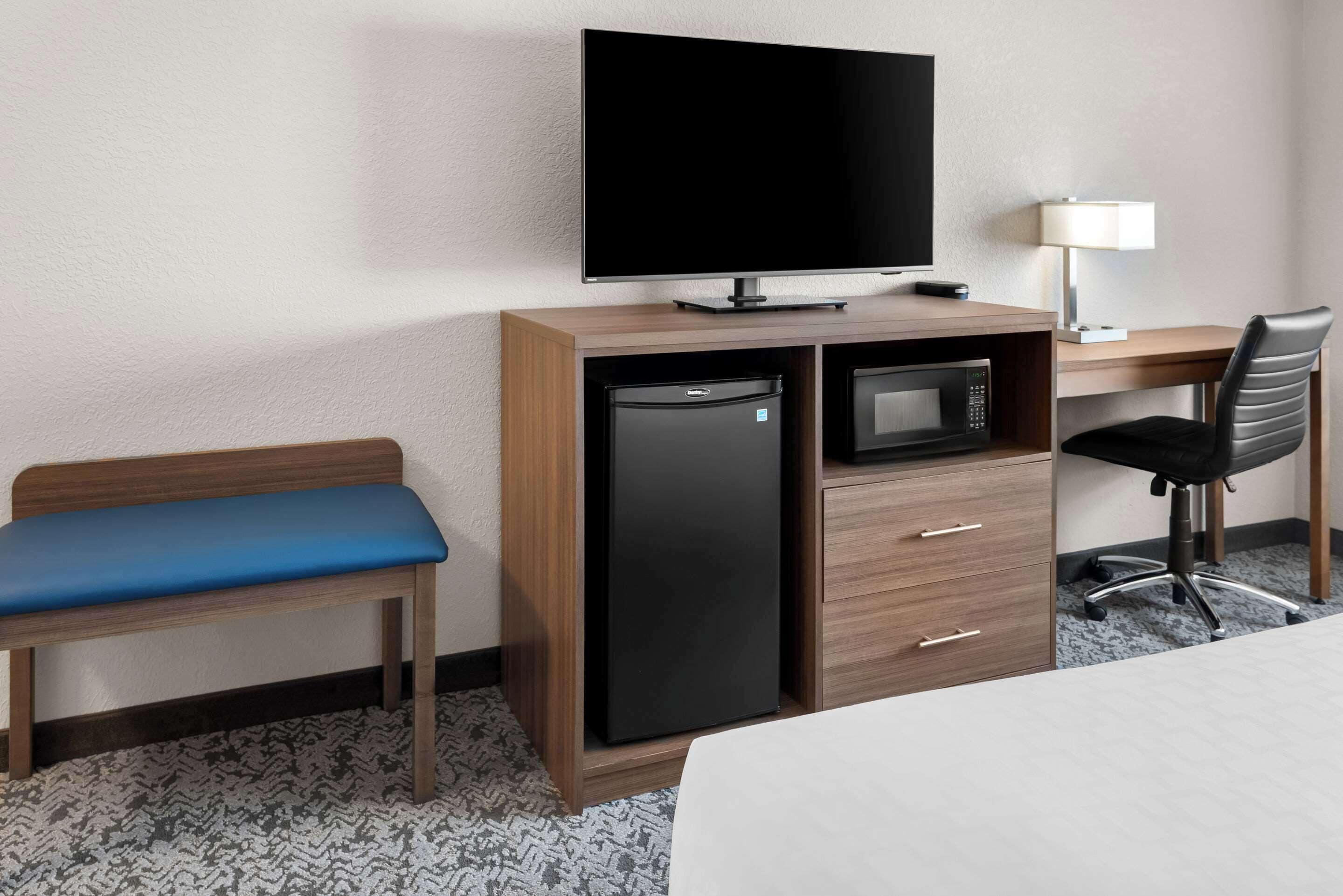Habitación de hotel con cama, escritorio con computadora, televisor, refrigerador, microondas, una silla y un banco.