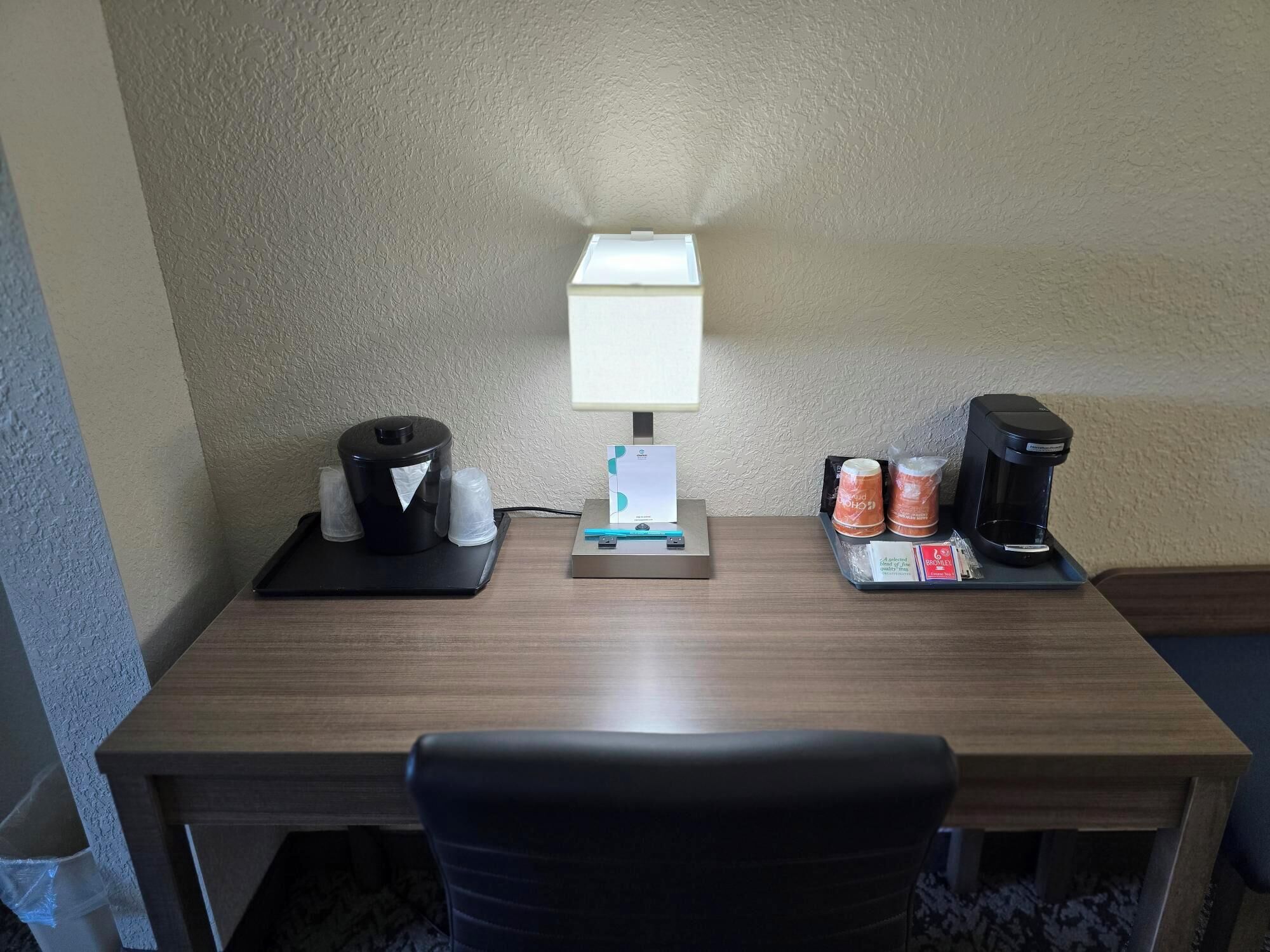 A hotel room desk with a lamp, a coffee maker, and a tray with coffee cups and a box.