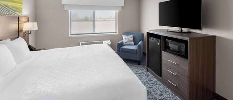 A hotel room with a large bed, a flat-screen TV mounted on a wooden cabinet, a small armchair, and a window with a view of a body of water.