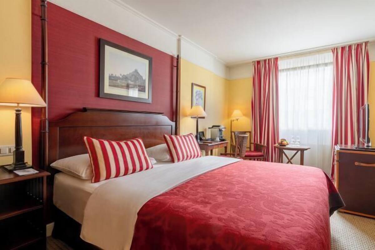 Een hotelkamer met een groot bed, een houten hoofdbord, twee nachtkastjes met lampen, een kleine tafel met een stoel, en een raam met gordijnen.
