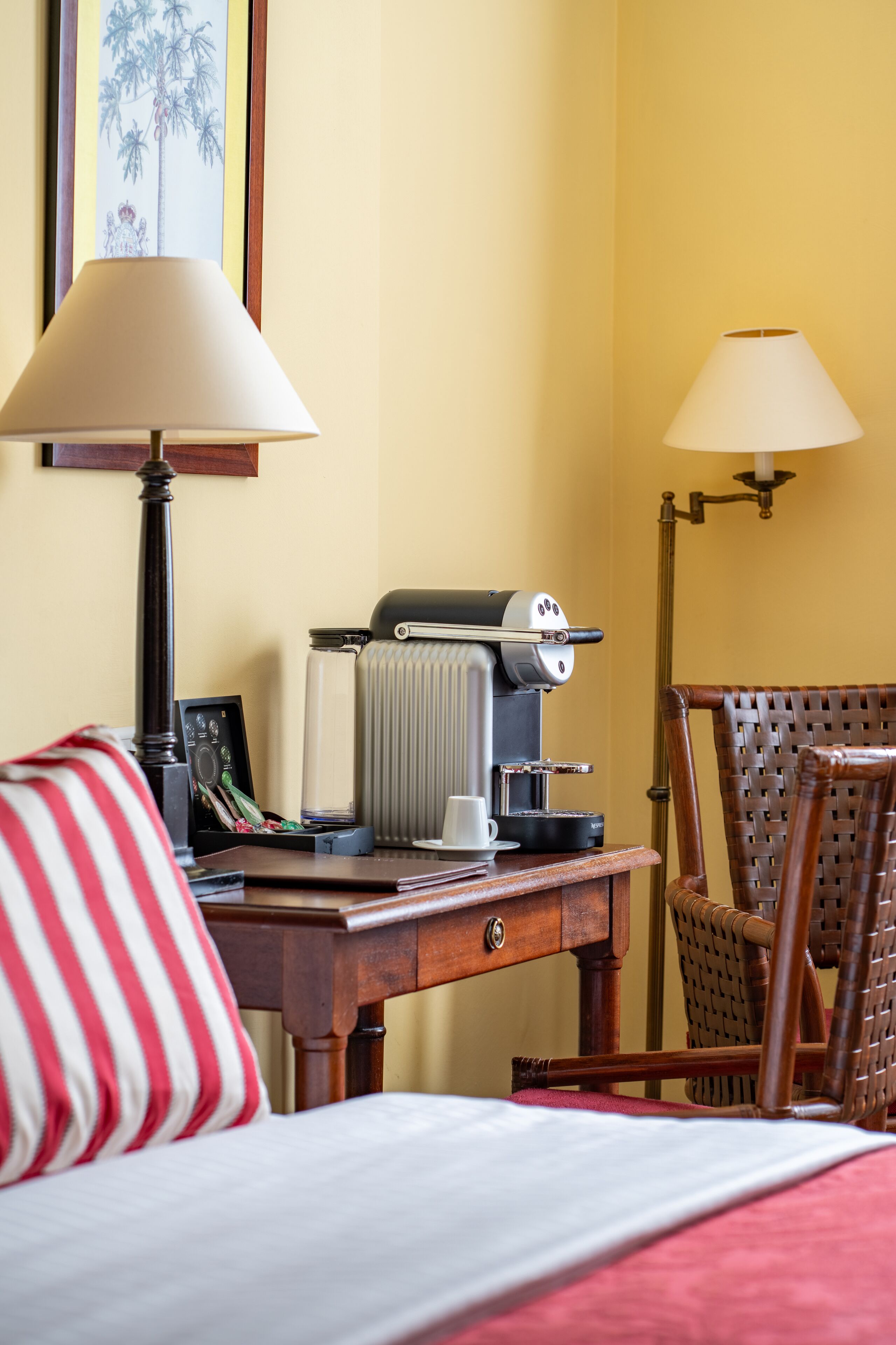 A hotel room with a bed, a wooden desk with a coffee maker, a lamp, and a wicker chair.