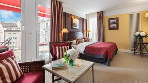 Habitación de hotel con una cama, una silla, una mesa con un libro y flores, y vistas a edificios a través de la ventana.