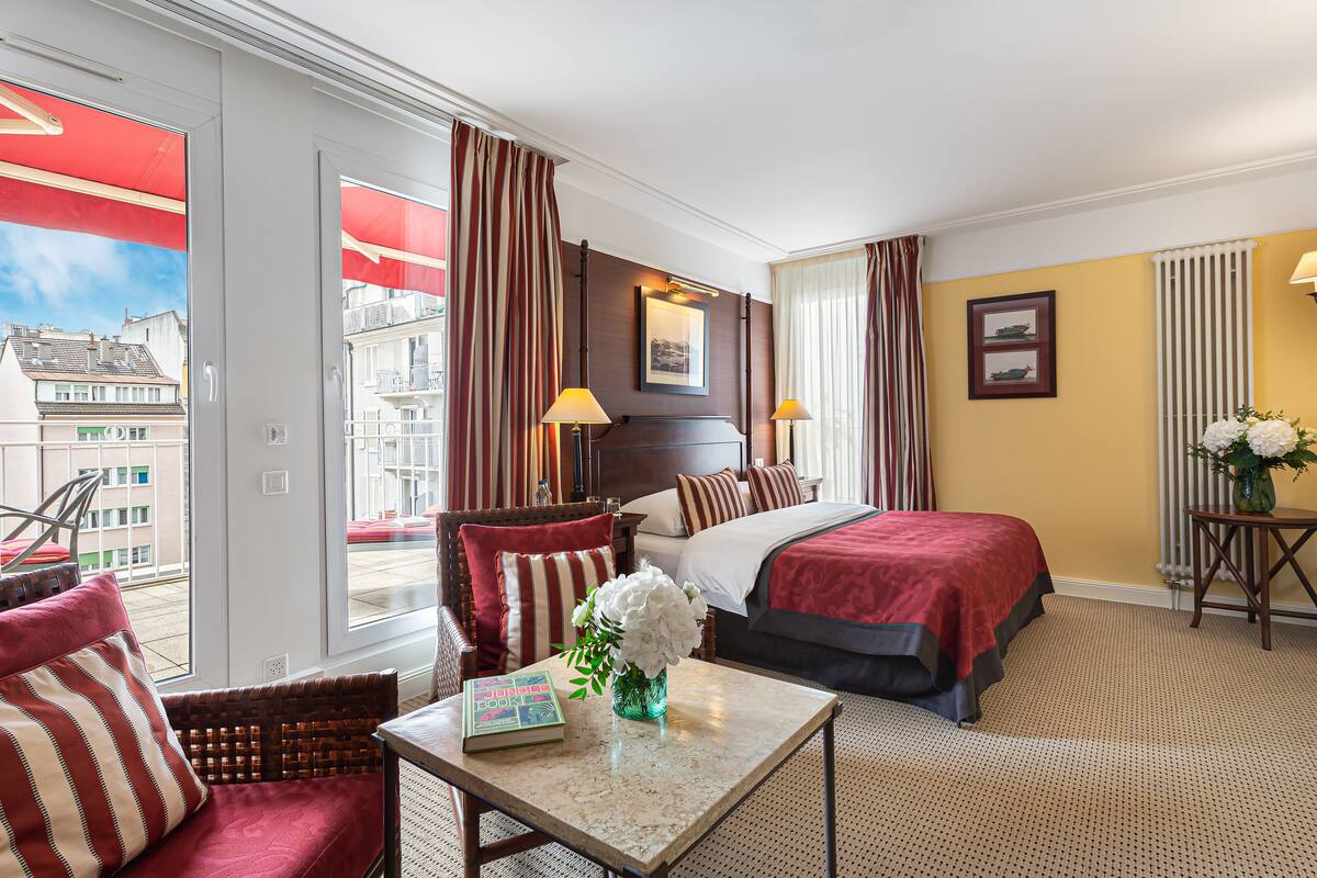 Een hotelkamer met een bed, een stoel, een tafel met een boek en bloemen, en uitzicht op gebouwen door het raam.