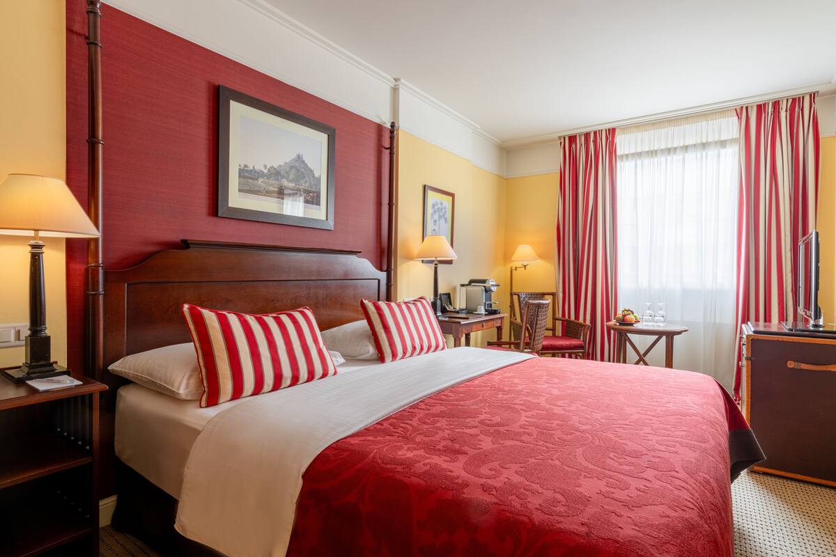 Een hotelkamer met een groot bed, twee nachtkastjes, een bureau, een stoel, een raam met gordijnen en een lijst met een afbeelding aan de muur.