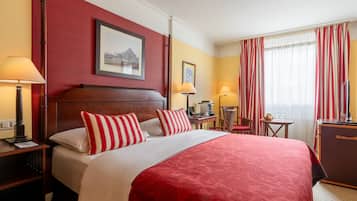 Een hotelkamer met een groot bed, twee nachtkastjes, een bureau, een stoel, een raam met gordijnen en een lijst met een afbeelding aan de muur.