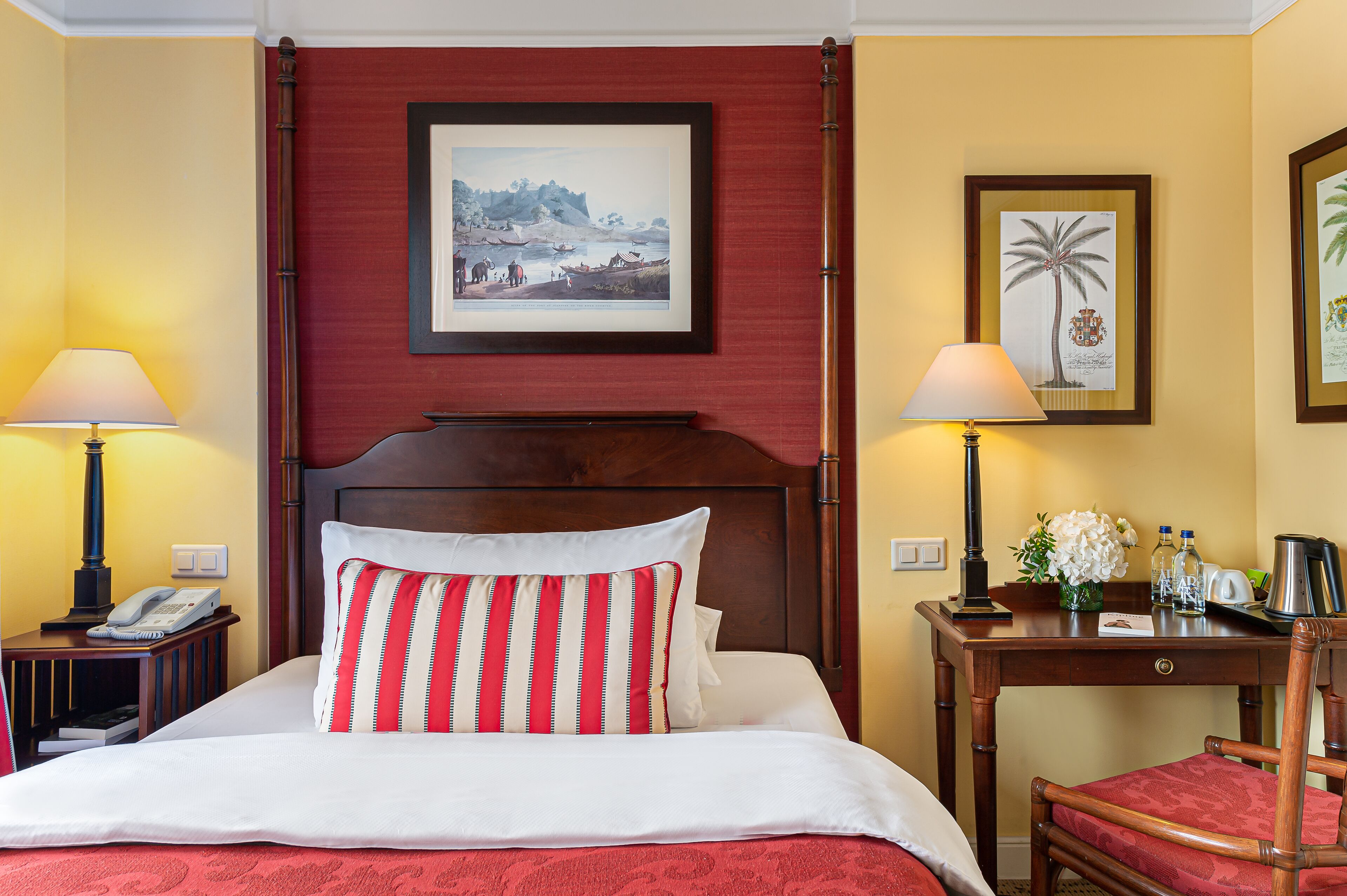 Habitación de hotel con una cama, dos lámparas de noche, una mesita de noche de madera con un teléfono, un cuadro en la pared, un escritorio con cafetera y una silla.