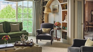 Un soggiorno elegante con un divano verde, una poltrona scura e un tavolino da caffè con libri e una teiera. Si può vedere una libreria piena di libri e oggetti decorativi, e una finestra che offre vista sul verde circostante.