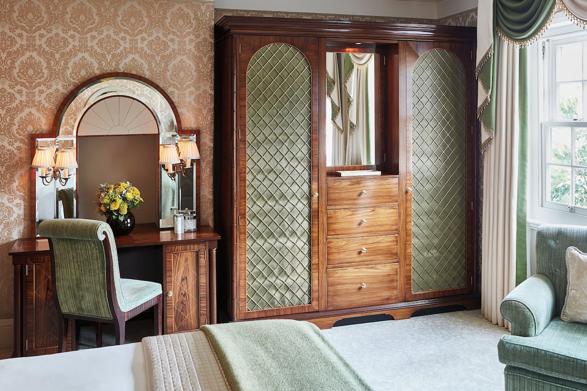 A bedroom with a large wooden wardrobe, a vanity with a mirror, a green upholstered chair, and a bed with a white comforter.