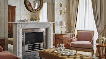 A classic room with a fireplace, a pink armchair, a tufted ottoman, and a mirror above the fireplace.