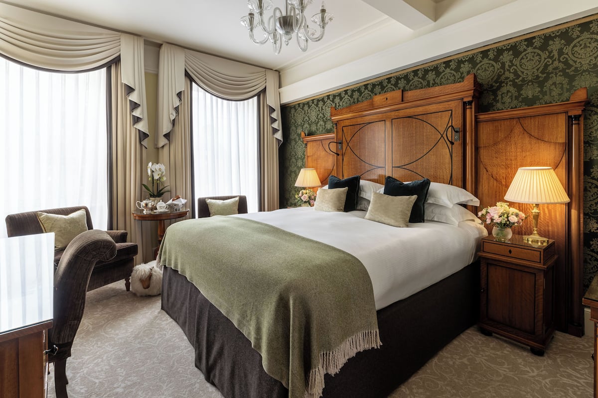 A spacious bedroom with a large bed, a wooden headboard, two bedside tables with lamps, a small table with a vase of flowers, and a chair.