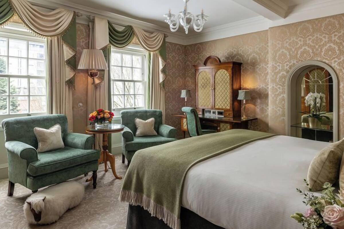 A classic bedroom with a large bed, two green armchairs, a wooden cabinet, and a small table with flowers.