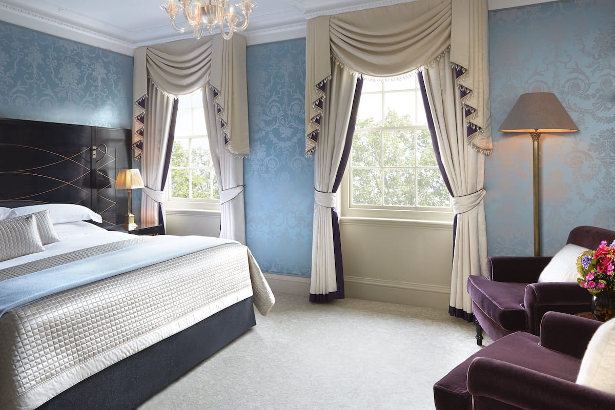 A hotel room with a large bed, two armchairs, a nightstand, a lamp, and a window with curtains.