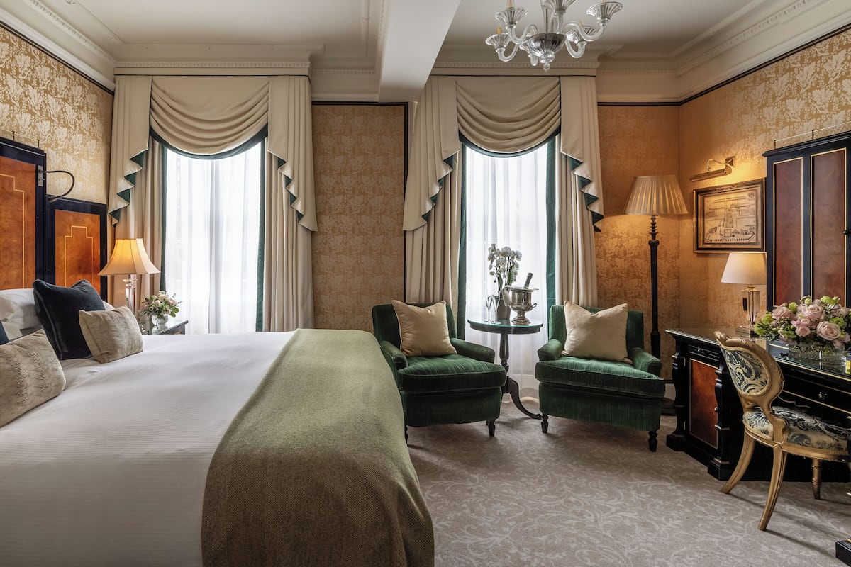 A hotel room with a large bed, two green armchairs, a small table with a lamp, and a decorative cabinet.