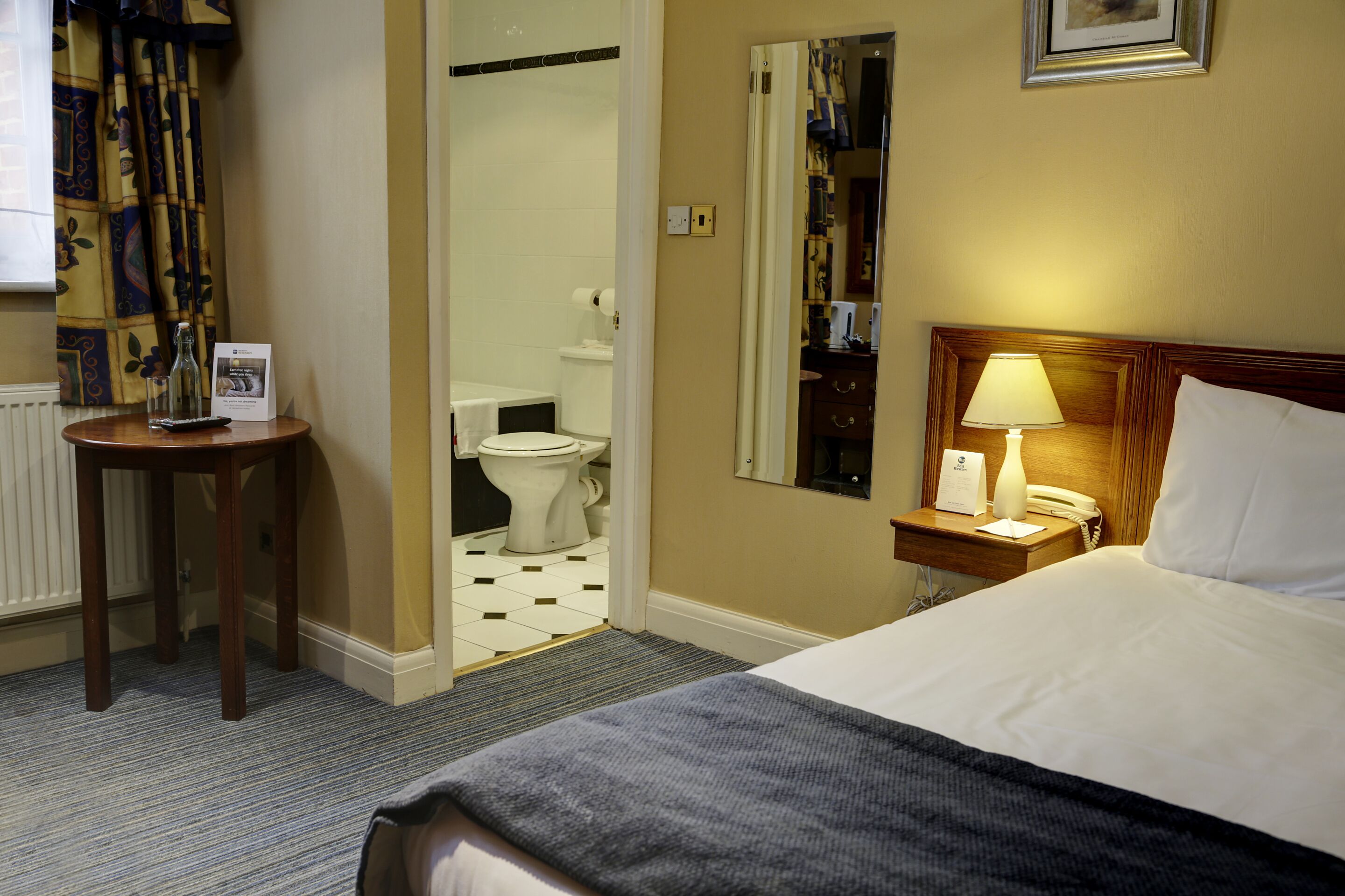 Habitación de hotel con cama, mesita de noche, lámpara y vista al baño.