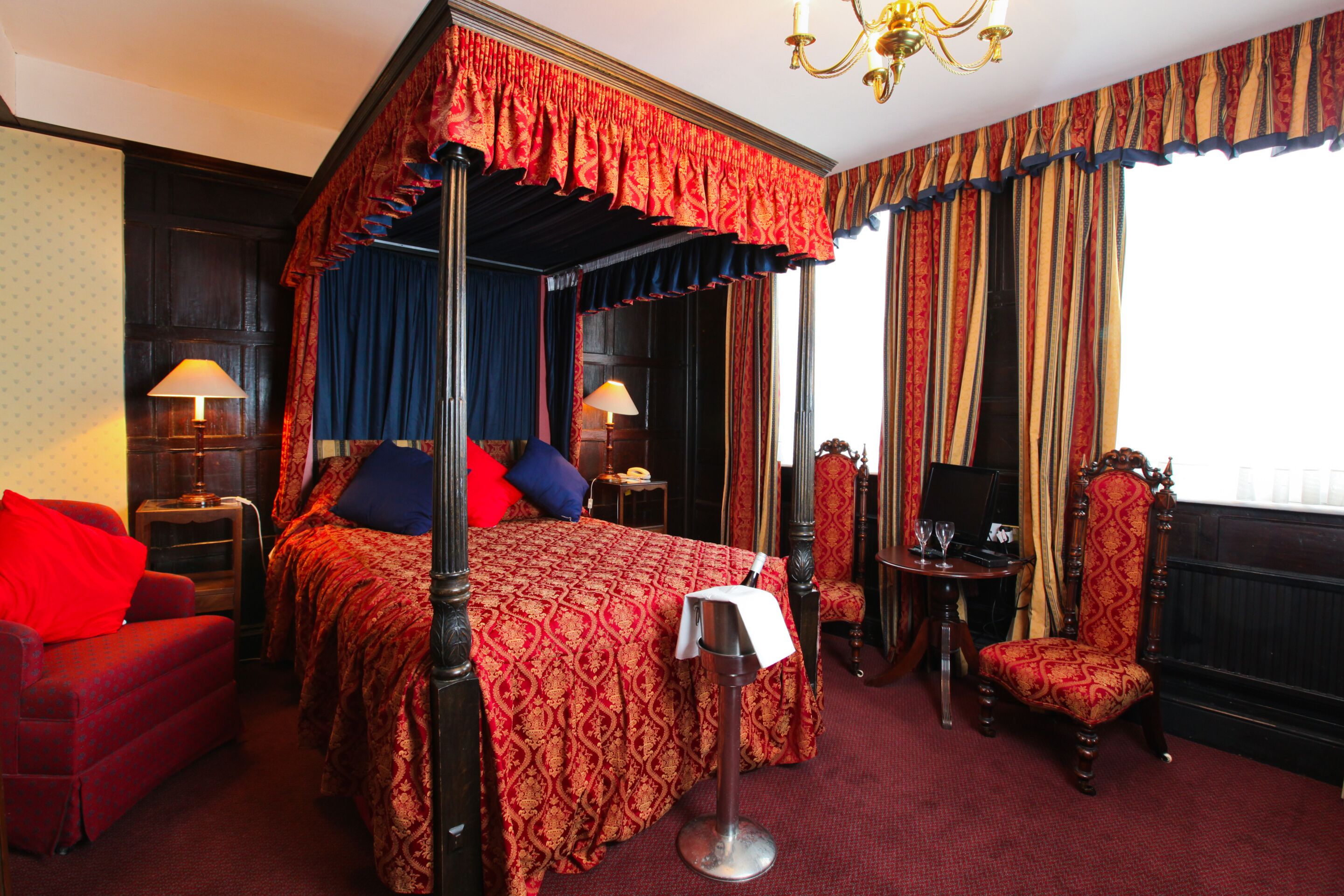 A four-poster bed with a canopy, a red upholstered chair, a desk with a lamp, and a television.