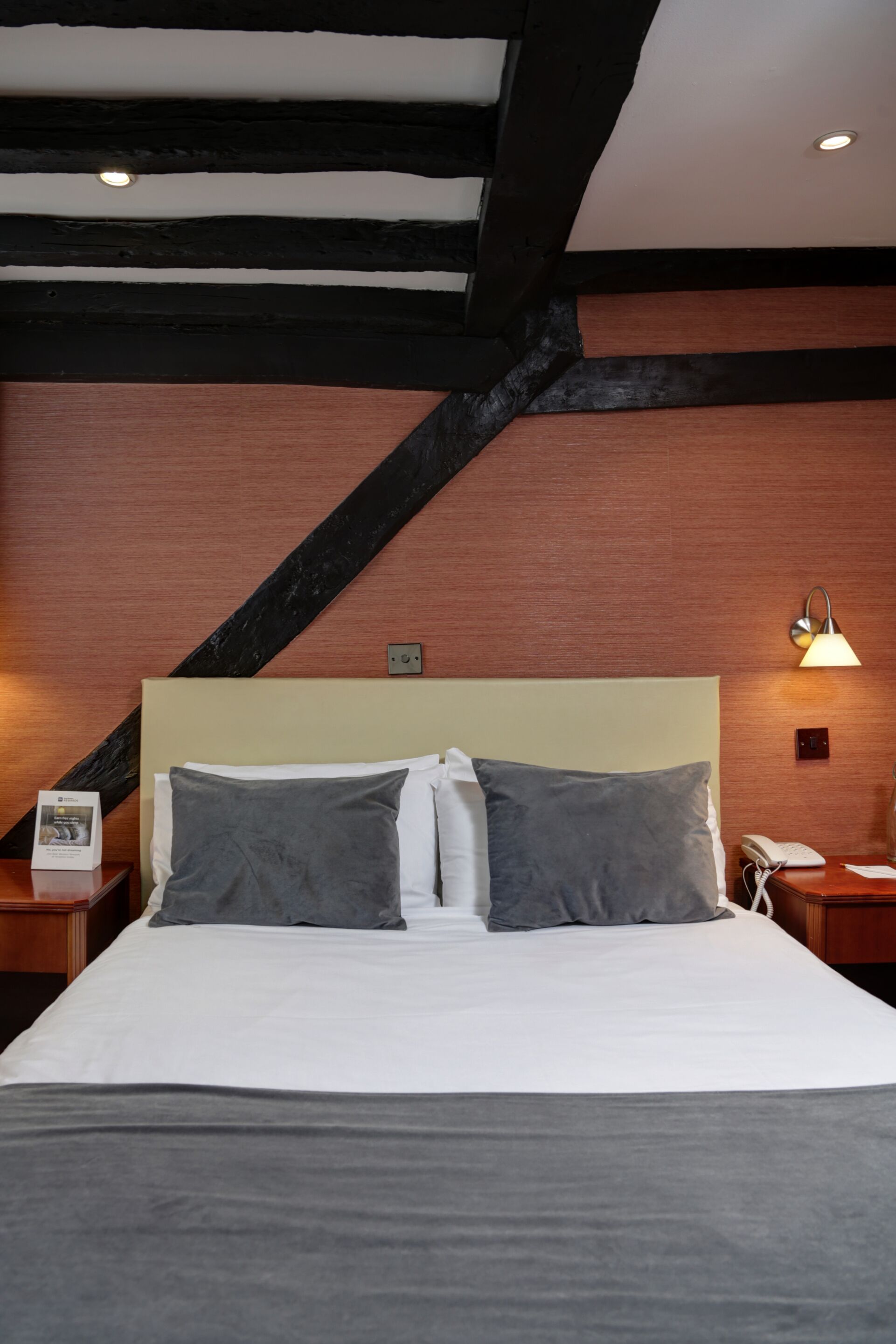 Habitación de hotel con una cama grande, vigas de madera, una mesita de noche con lámpara y un teléfono en la pared.